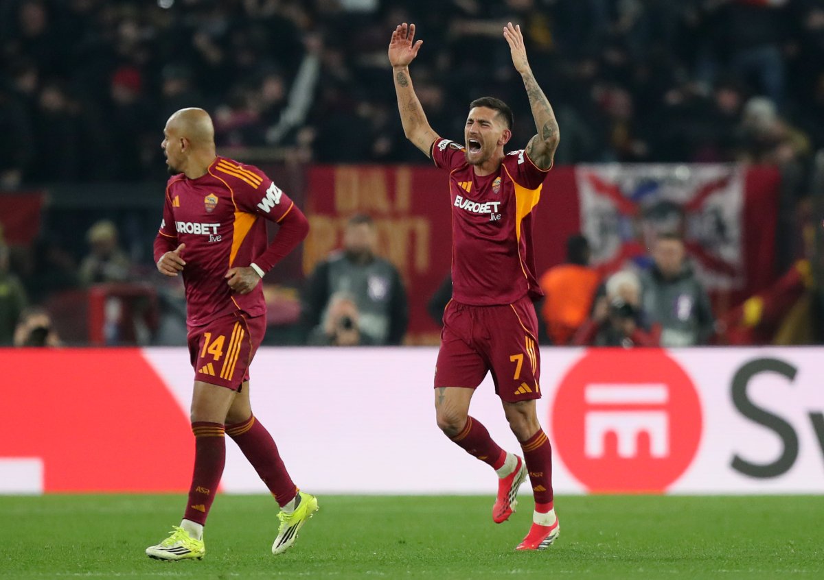 Two AS Roma players celebrate a goal on the pitch with arms raised, stadium crowd in the background.