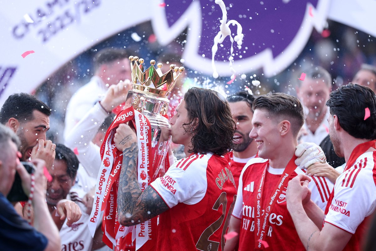 Soccer player kisses the Premier League trophy as teammates celebrate with ribbons and confetti.