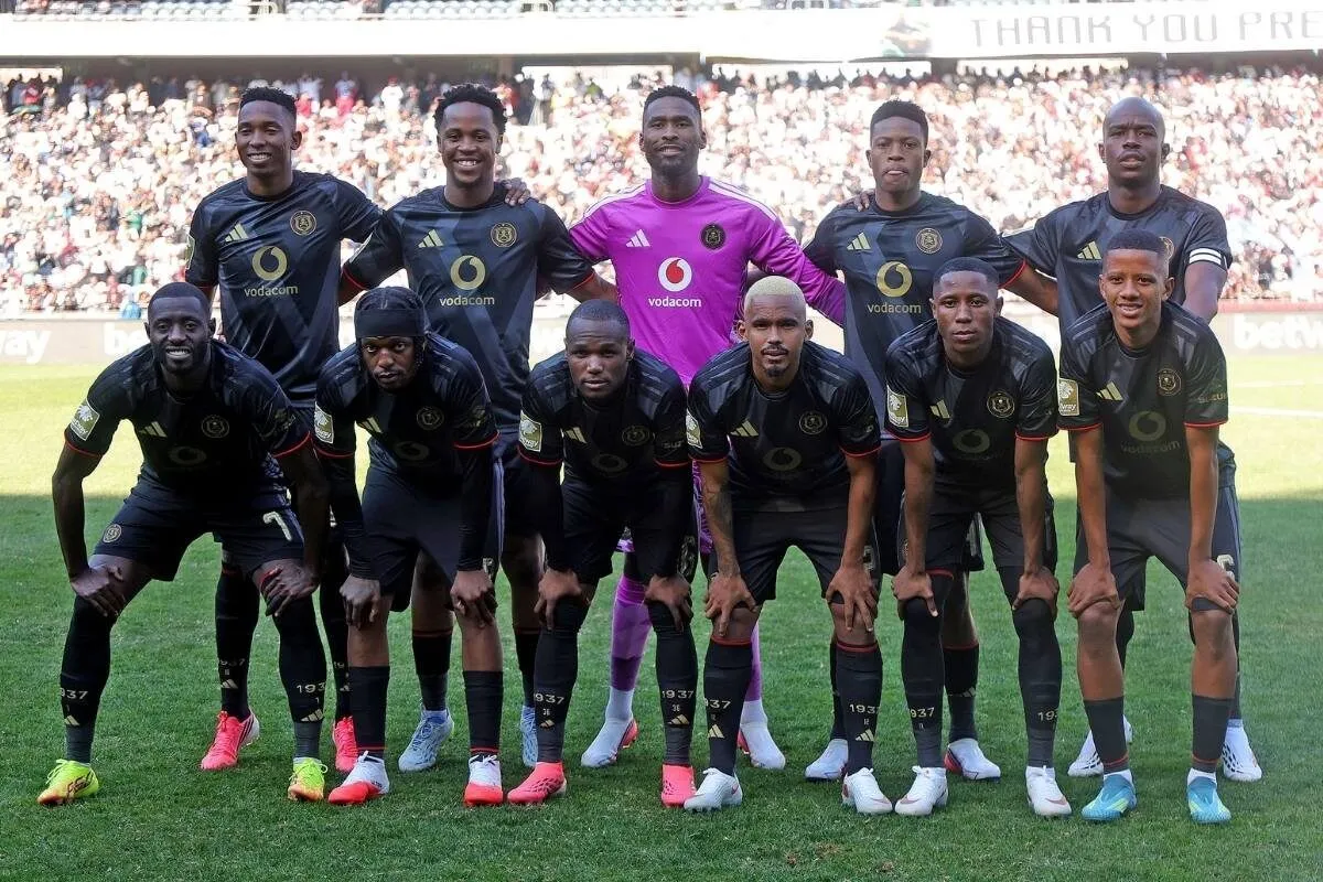 Group of ten soccer players in black uniforms pose for a team photo on a green pitch, with a purple-clad goalkeeper in the back row center and a crowded stadium behind them.