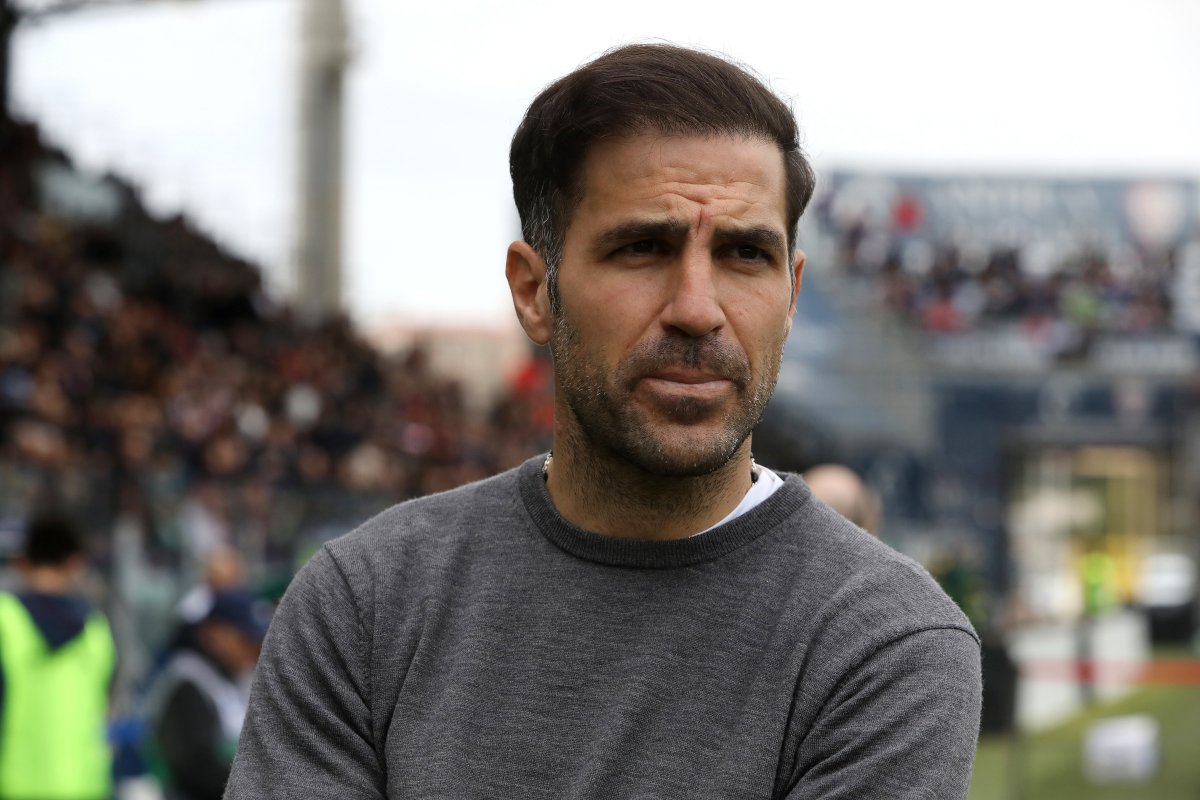 Man with short dark hair and stubble wearing a gray sweater, standing outdoors at a crowded event, looking contemplative.