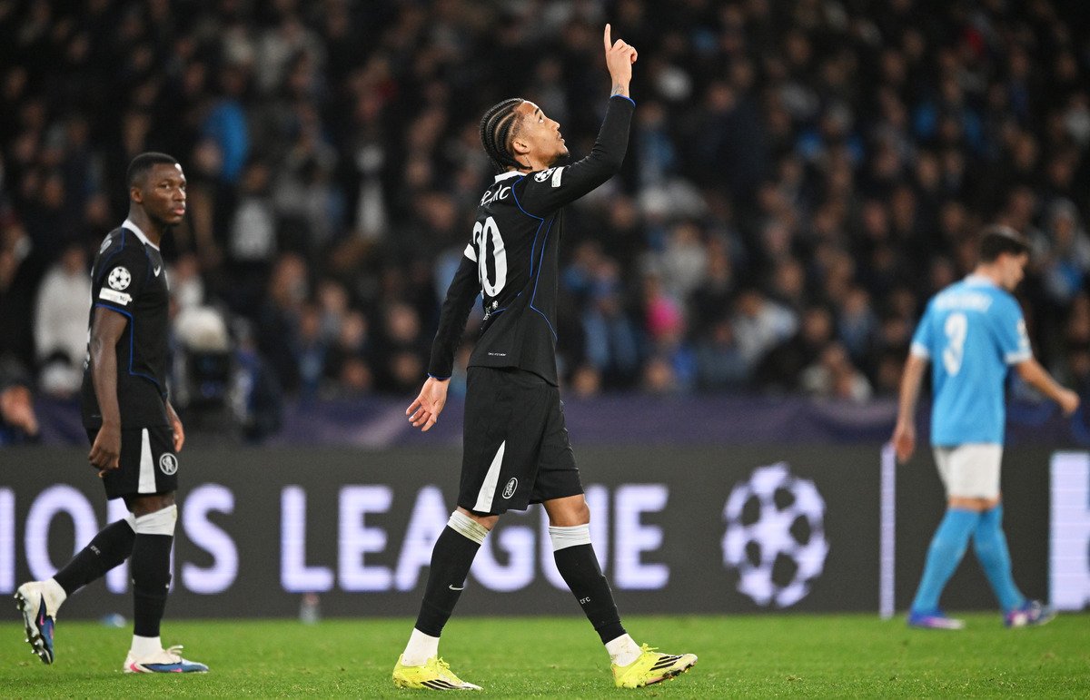 Footballer in a black kit with number 20 points upward while celebrating on the field; crowd in the stands behind him and a blue-clad opponent in the background.