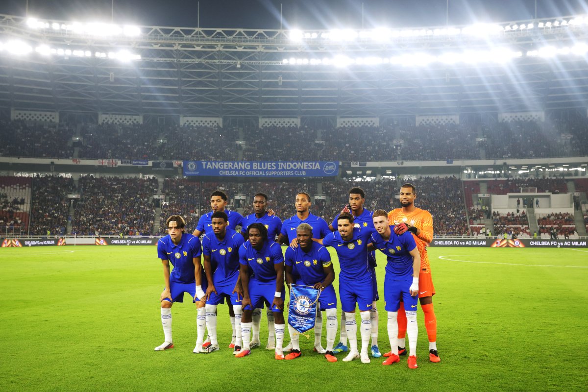 Chelsea FC players in blue uniform pose for a group photo on the stadium pitch with a packed crowd behind them.
