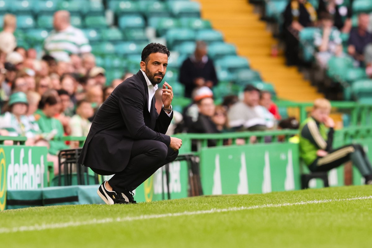 Man in a black suit crouches at the sideline of a green soccer field, watching the game amid stadium seats.