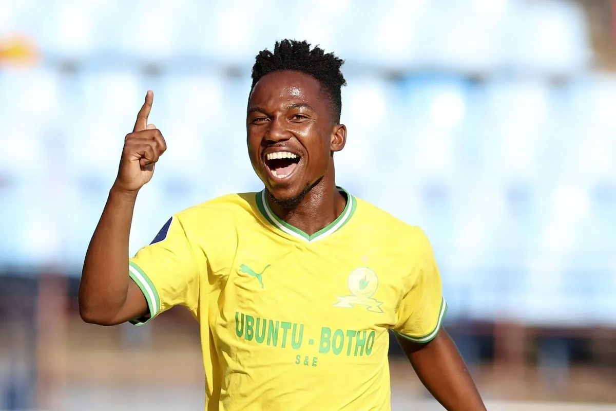 Soccer player in a yellow jersey with green trim celebrating, pointing upward with a wide smile on a sunny field.