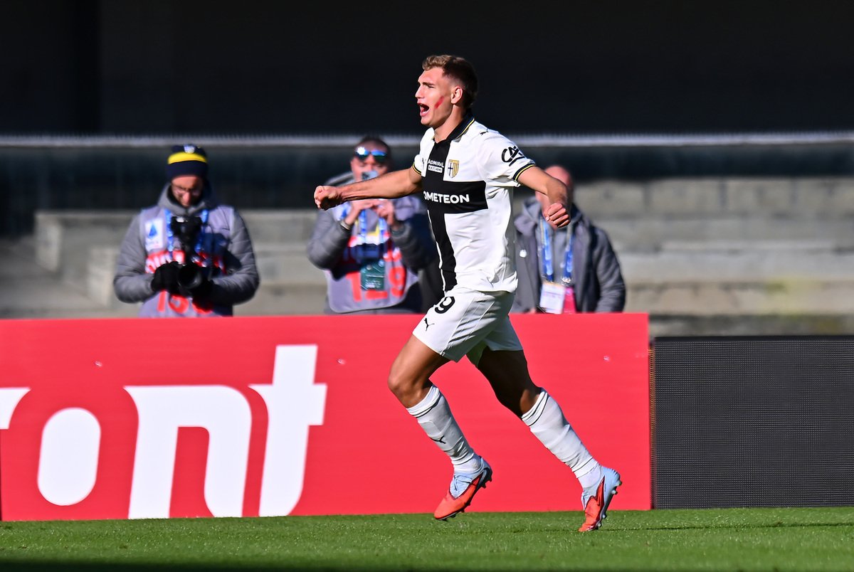 Soccer player in a white kit celebrates a goal, running on the field with photographers in the background.