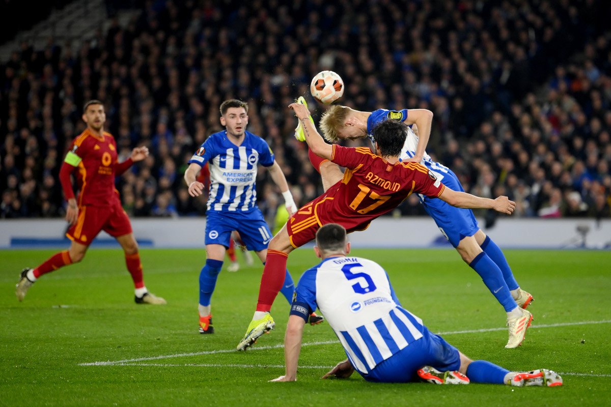 Action-packed soccer moment: a red-team player leaps as a blue-and-white defender challenges, ball in midair amid a crowded stadium.