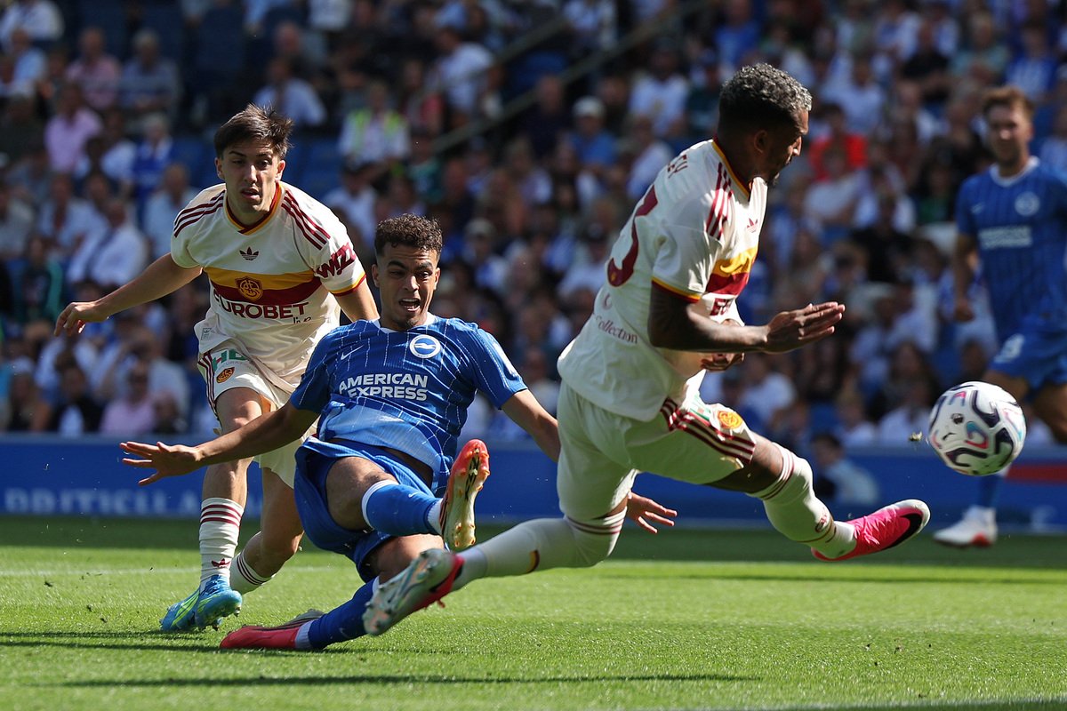 Soccer match moment: a blue-kit player slides in as two white-kit players approach, with the ball near the right edge of the frame and a crowd behind.