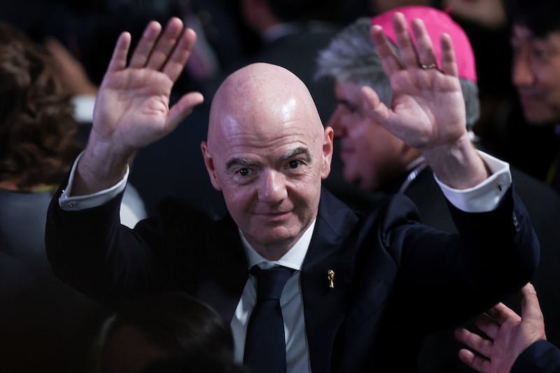 Bald man in a dark suit and blue tie raises both hands in a greeting at a formal event.