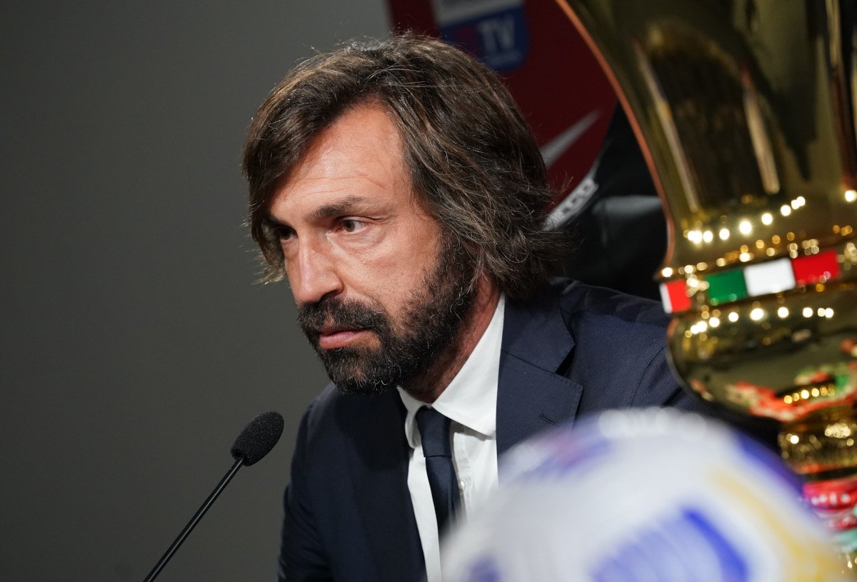 Man in a dark suit and white shirt speaks into a microphone during a press conference, with a large gold trophy partially visible beside him.