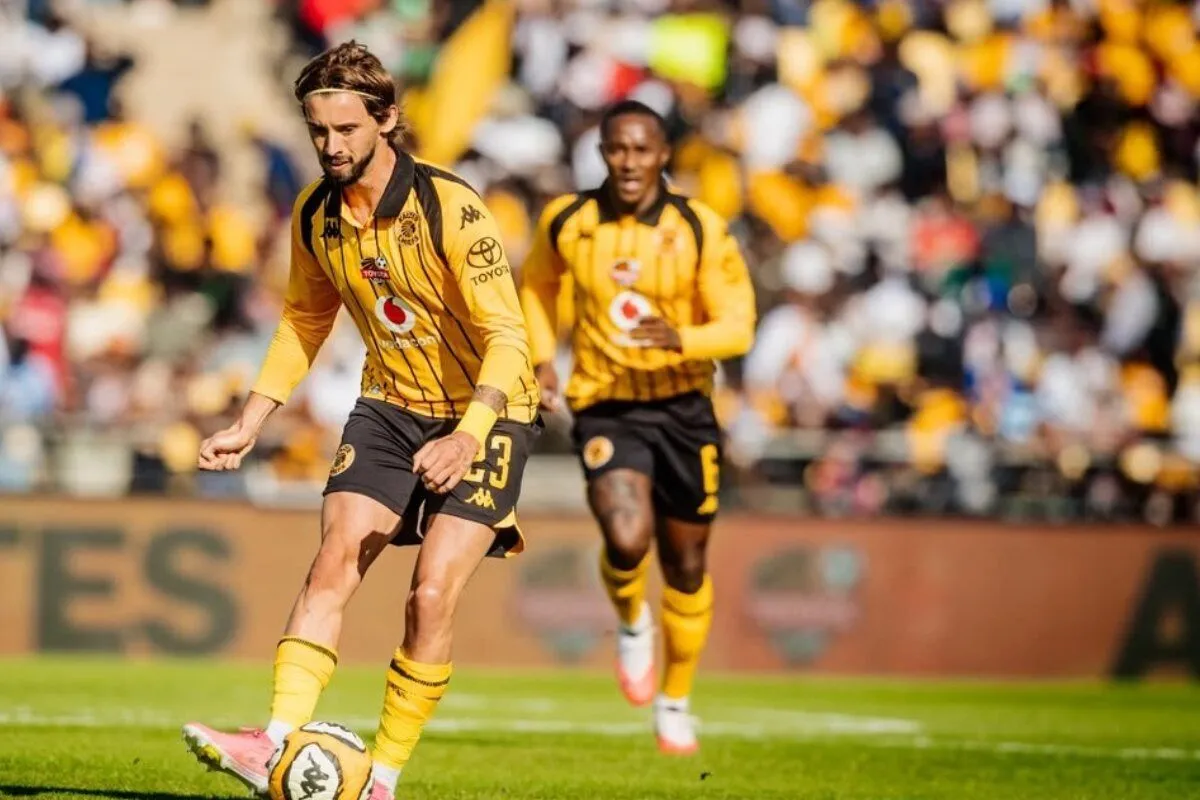 Soccer player in yellow and black kit controls the ball on a green field with a crowd watching in stadium behind him.