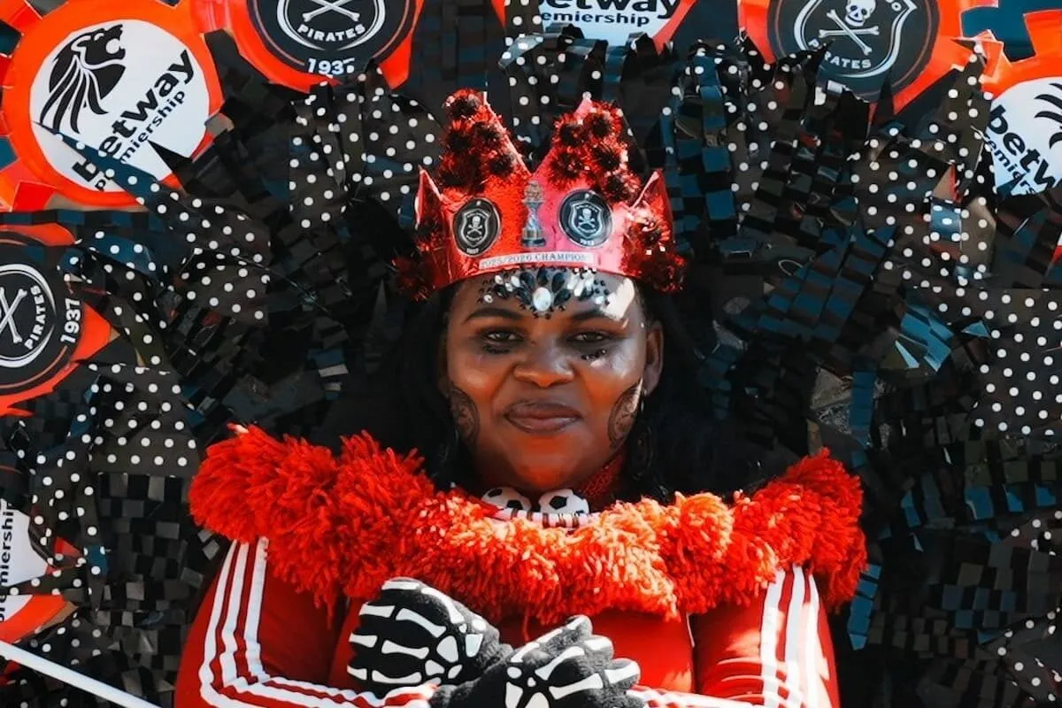 Person in a red carnival costume with a feathered collar, red crown, and skeletal gloves, posing for a photo on a parade backdrop of black streamers and dots. Person in a red carnival costume with a feathered collar, red crown, and skeletal gloves, posing for a photo on a parade backdrop of black streamers and dots.