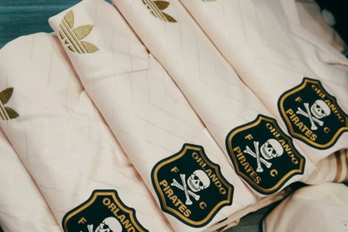 Close-up of pale pink sports jerseys with gold Adidas logos and Orlando Pirates crests showing a skull and crossbones comic.