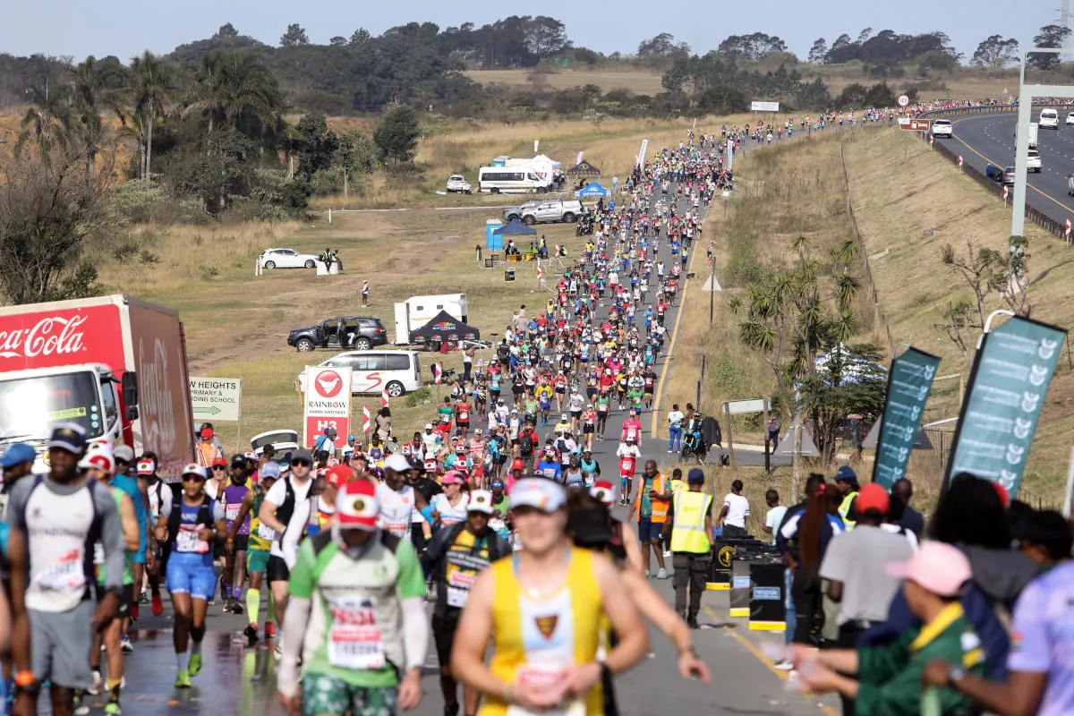A crowded road marathon with thousands of runners, along a highway lined by tents, volunteers, and support vehicles on the side.