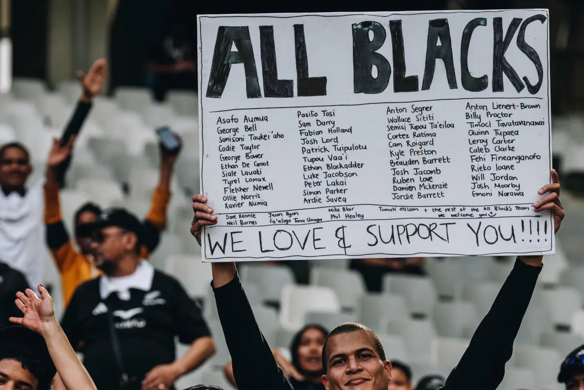Supporters in a stadium hold a large handwritten banner reading 'ALL BLACKS' and 'WE LOVE & SUPPORT YOU!!!!'