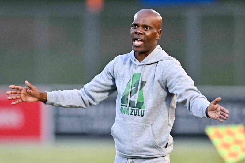 A man in a gray hoodie with a green logo stands on a sports field, arms spread wide as if giving instructions or arguing playfully.