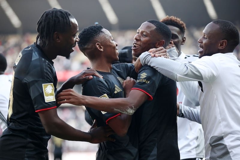 Soccer players in black uniforms celebrate with a group hug on the field, stadium in the background.