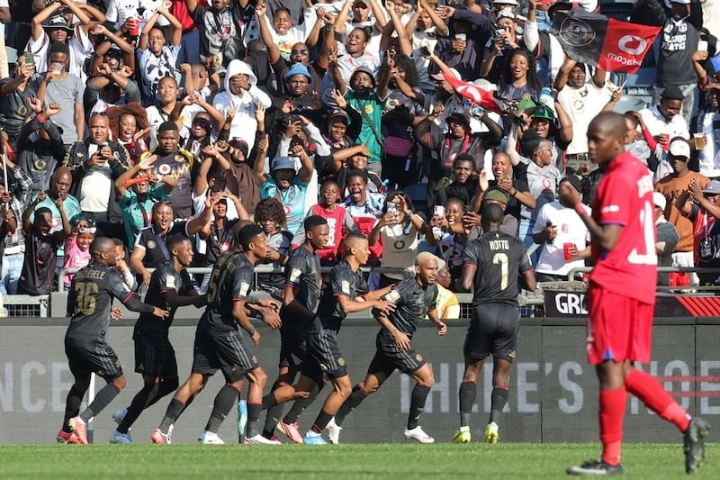 Soccer players in black kits celebrate a goal as teammates run toward the cheering crowd; a player in red stands to the right.