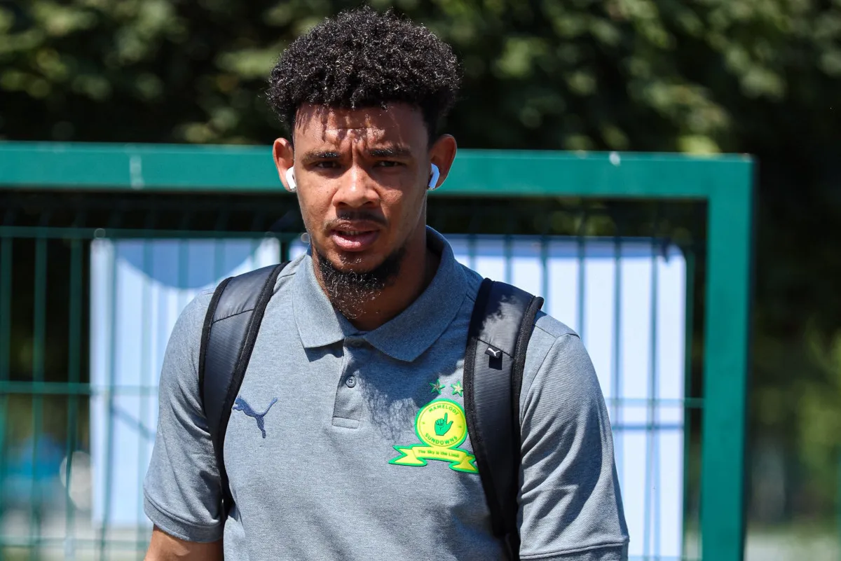 Young man wearing a gray polo shirt with a green-and-yellow team badge, backpack, and wireless earbuds, outdoors by a green fence.