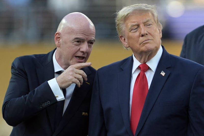 Bald man in a dark suit points at a second man in a navy suit with a red tie at an outdoor event.