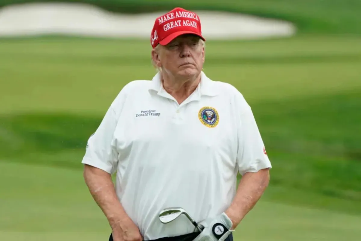 Man on a golf course wearing a red MAGA hat and white polo, holding a golf club.