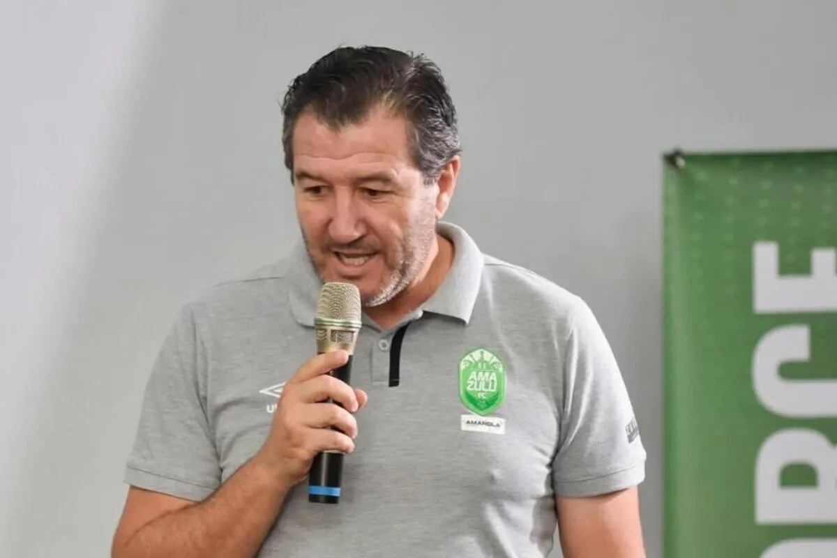 Man in a gray polo shirt speaks into a handheld microphone at an indoor event, wearing a team crest on his chest.