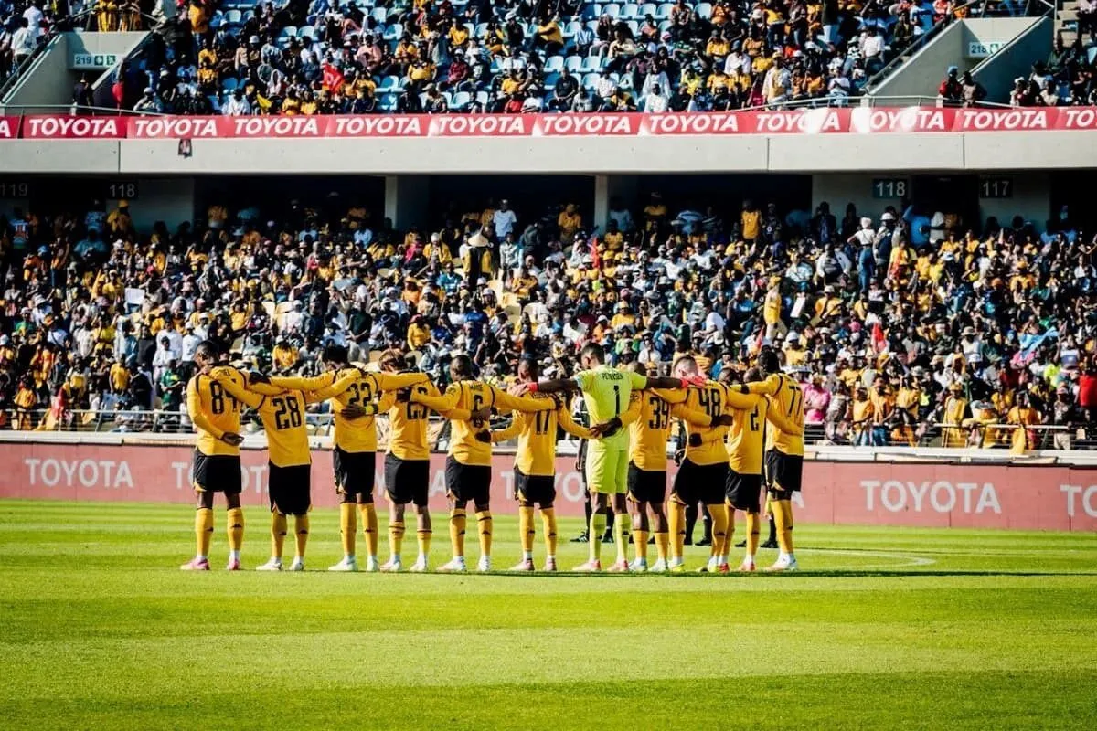 Soccer players in yellow jerseys stand in a line with arms around each other on a sunny field, a cheering crowd in the stands behind them.