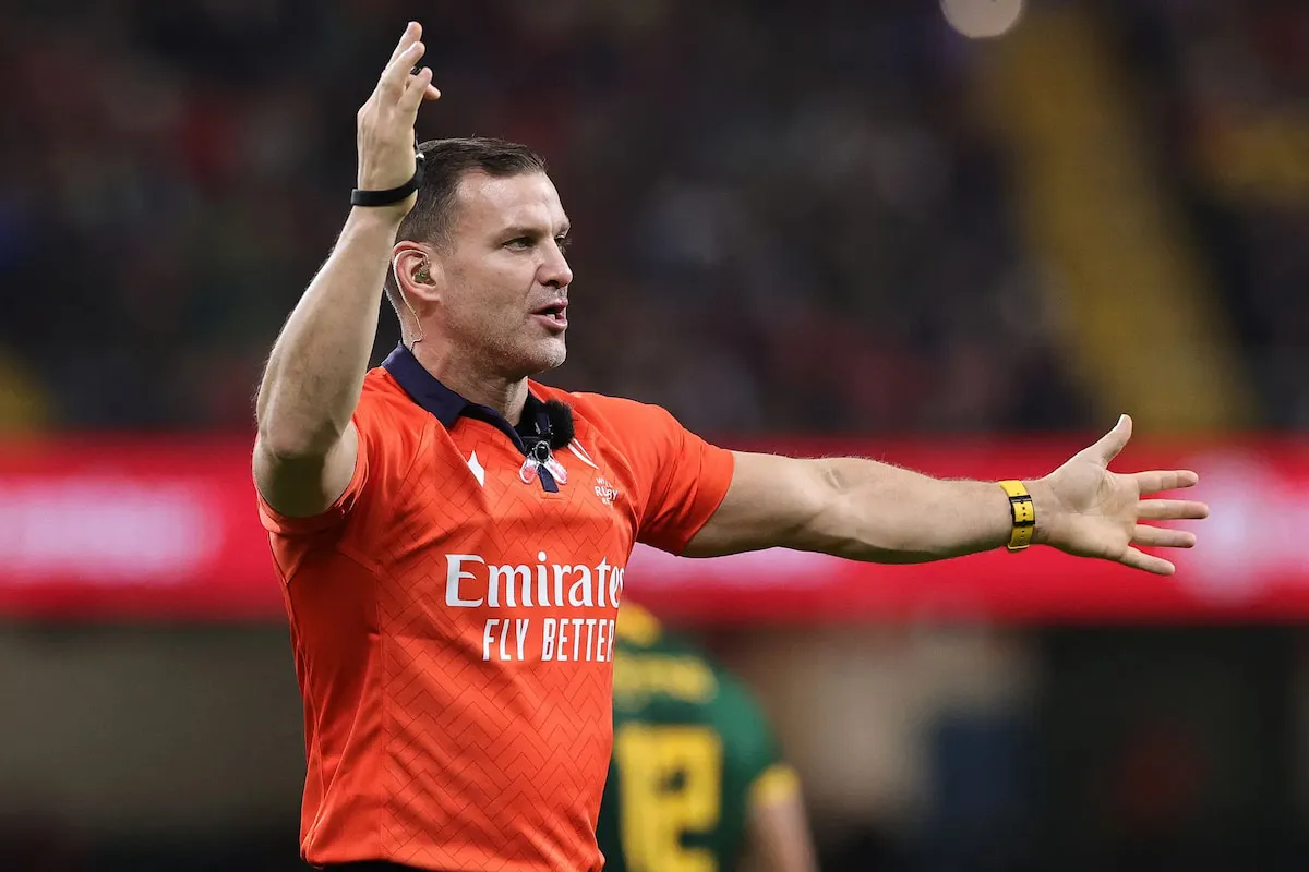 Rugby referee in an orange shirt raises both arms on the field, signaling a call with the Emirates Fly Better logo visible.