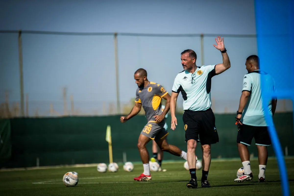 Soccer coach in a light blue shirt raises a hand while guiding players during a training session on a green field with balls nearby.