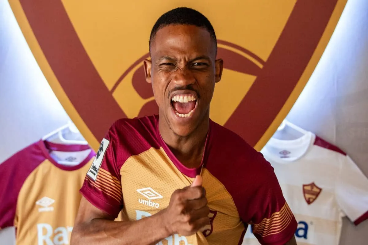 Young man in a maroon and gold football jersey cheers with mouth open, giving a thumbs up in front of a team crest background.