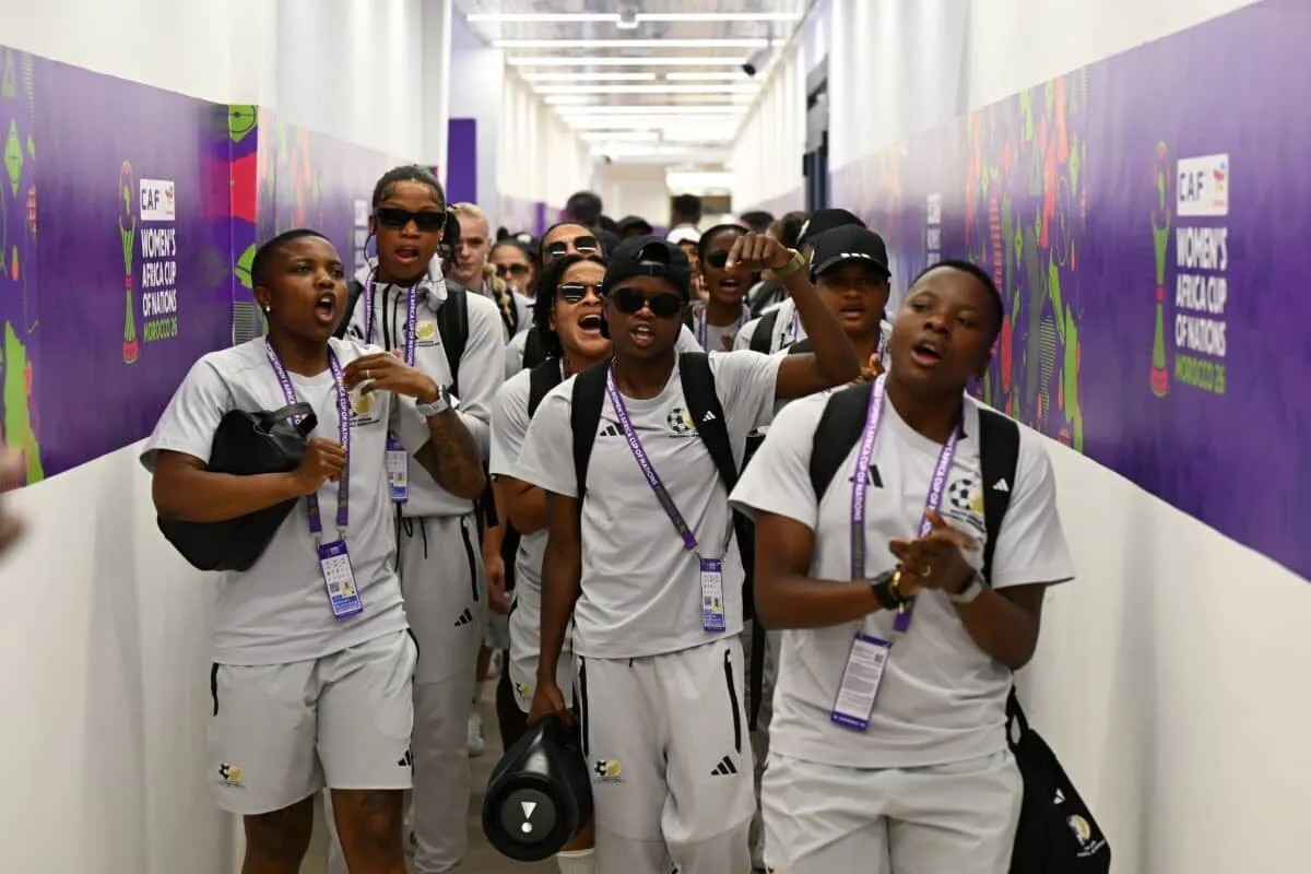 Group of young soccer players in white uniforms walk down a purple-lit corridor, cheering with lanyards and backpacks.