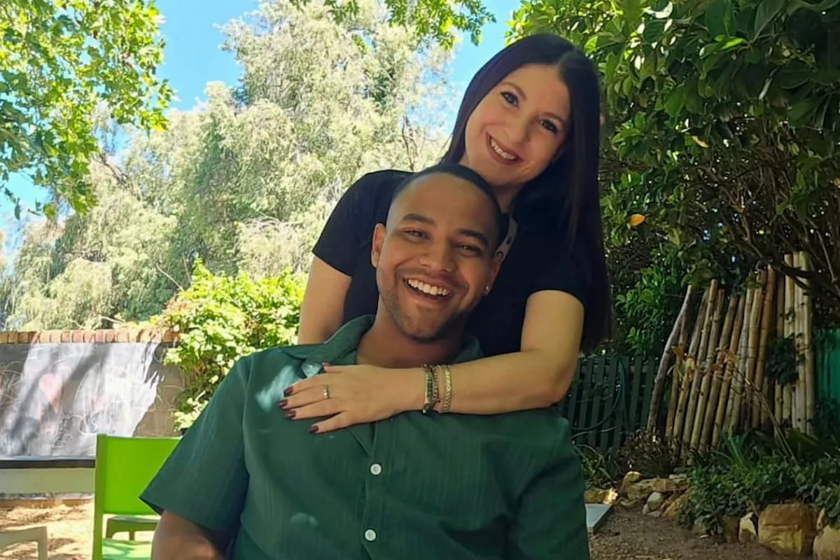 Smiling woman stands behind a smiling man, arms around his shoulders in a sunny garden.