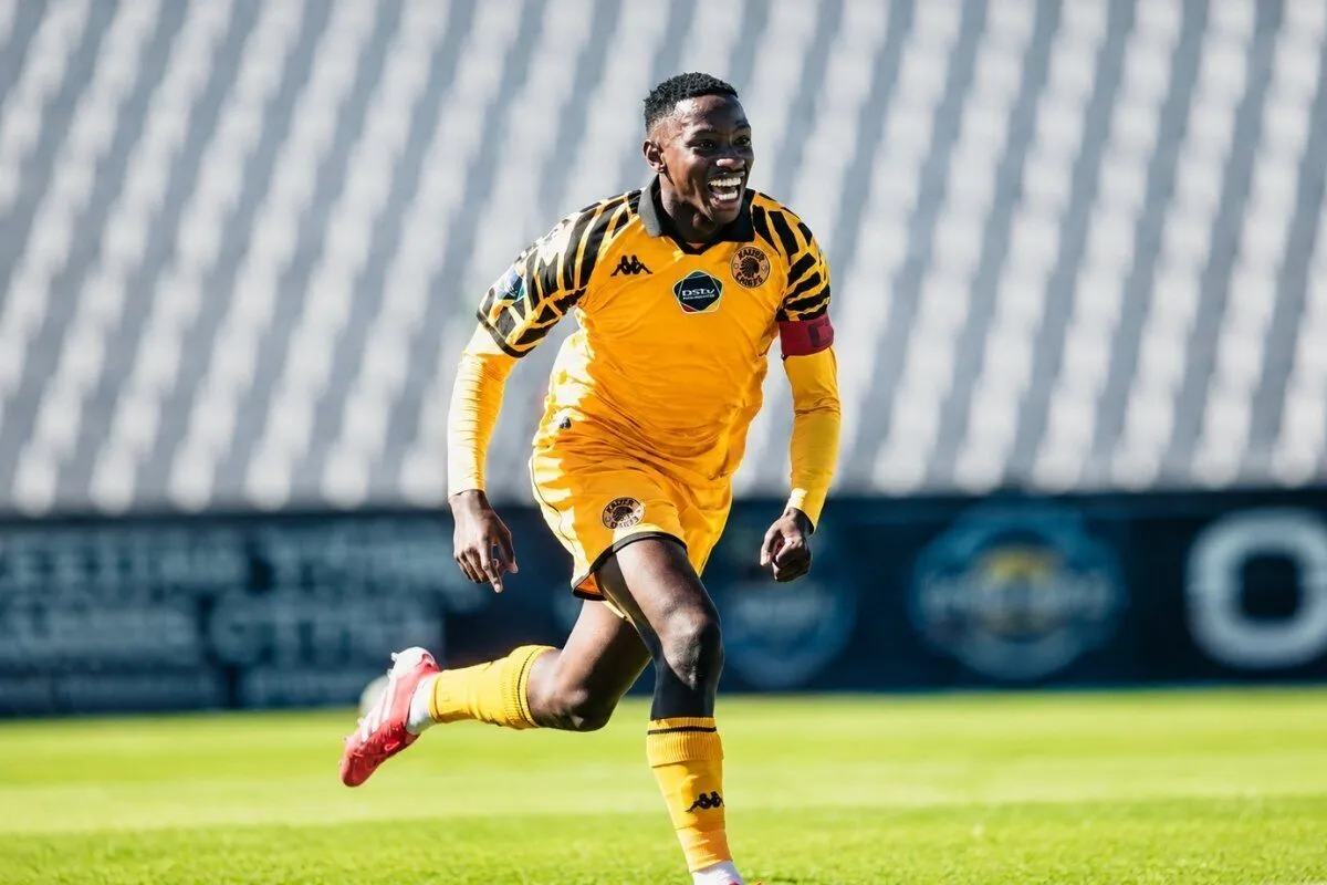 Soccer player in a yellow kit running on a green field with stadium stands in the background, celebrating or in action.