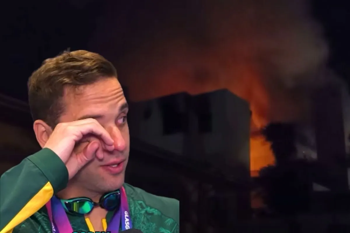 Man in a green jacket wipes his eye as a burning building smolders in the background.