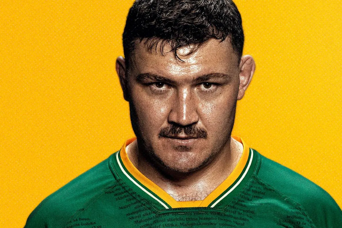 Close-up of a man with a stern expression, wearing a green sports jersey with yellow trim against a bright yellow background.