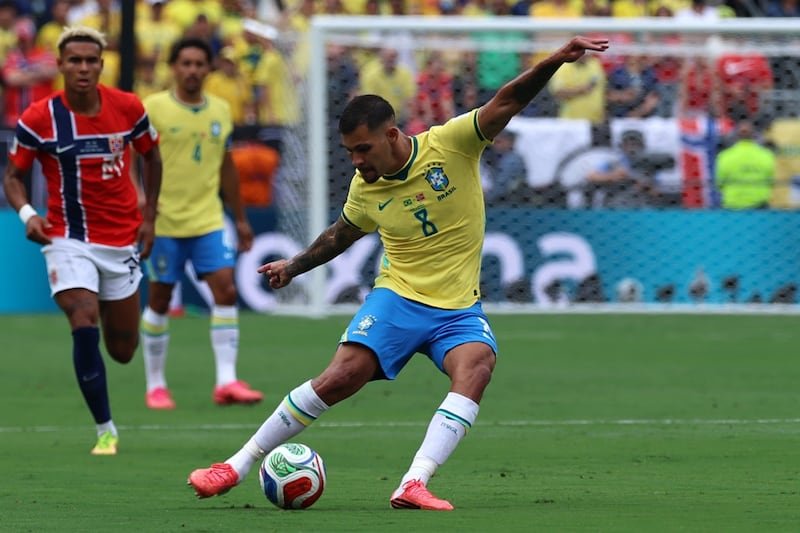 Brazilian soccer player in yellow jersey (number 8) kicking a ball on the field during a match, with teammates and goal in the background.