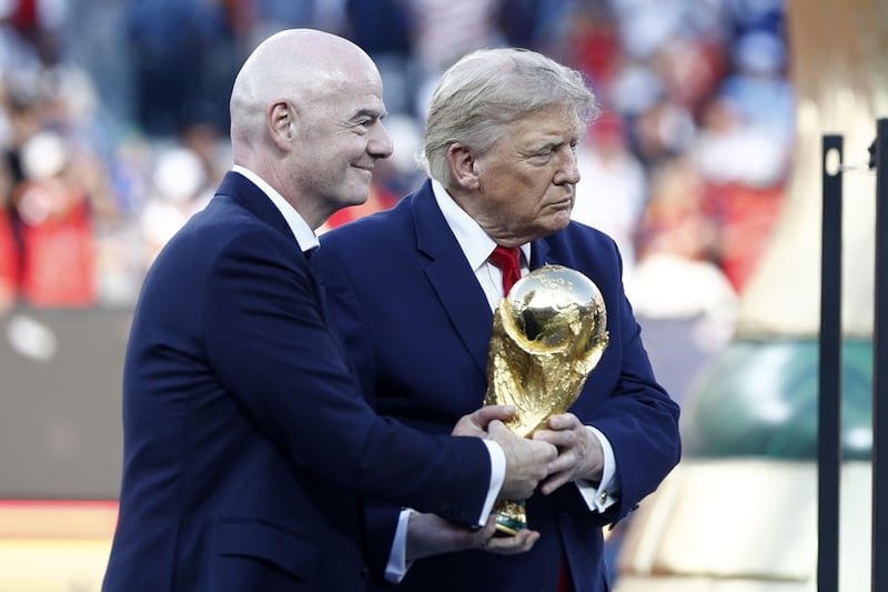 Two men in dark suits hold a large gold trophy during a ceremony on a stadium field.