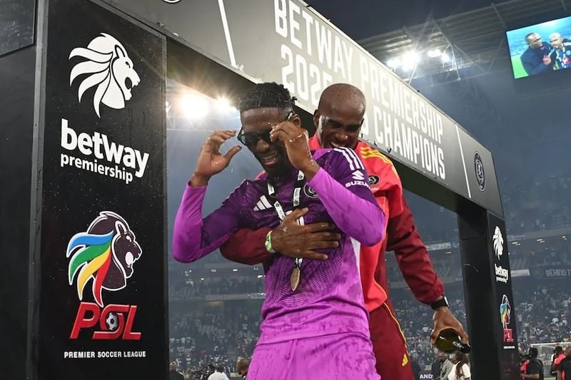 Two soccer players celebrate on a champions podium, one in purple with a medal being hugged by a teammate in red; Betway Premier League signage in the background.