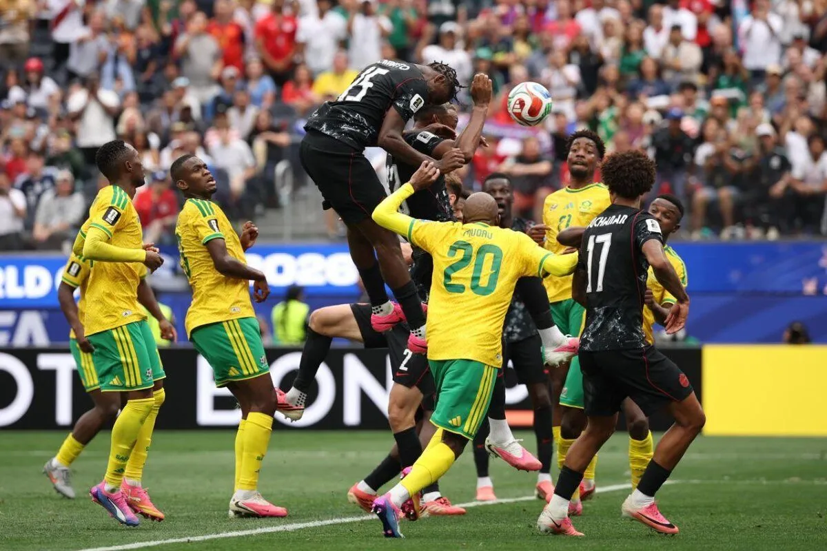 Soccer players from yellow-green and black teams jump to head the ball during a match with a crowded stadium in the background.