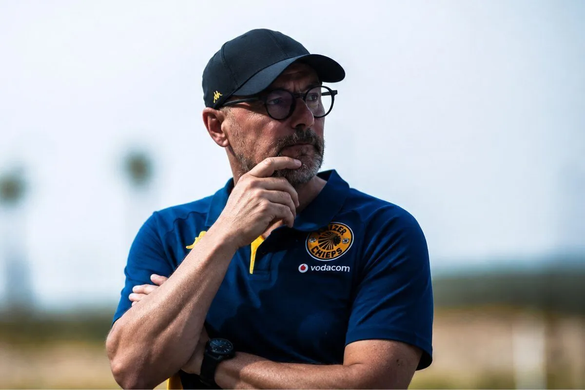Man in blue polo with Chiefs logo and Vodacom sponsor, wearing a cap and glasses, chin resting on his hand outdoors.