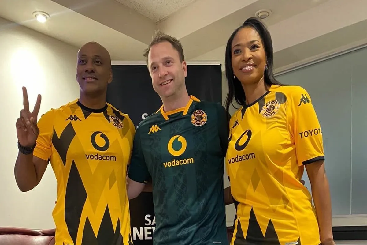 Three people pose indoors, two in yellow Kaizer Chiefs jerseys and one in a green goalie jersey, all smiling with vodacom logos visible on the shirts.