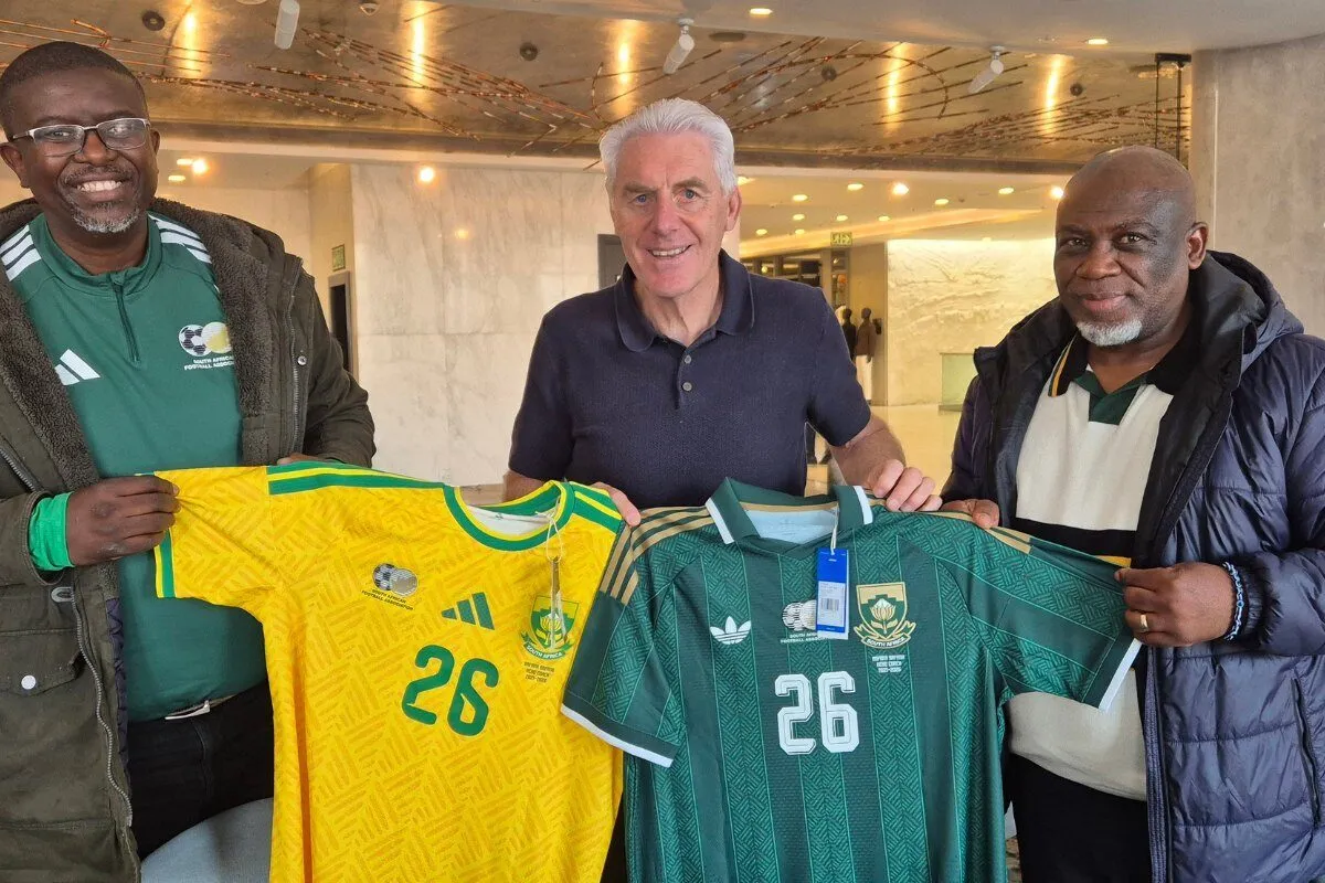 Three men pose in a bright lobby, holding up two soccer jerseys: a yellow kit with green trim and a green kit with white accents, both numbered 26, with badges on the chest area.