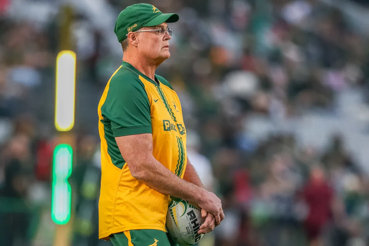 Older man in green and gold rugby kit holding a ball, standing on the sideline with stadium and blurred crowd in the background; wearing cap and glasses.