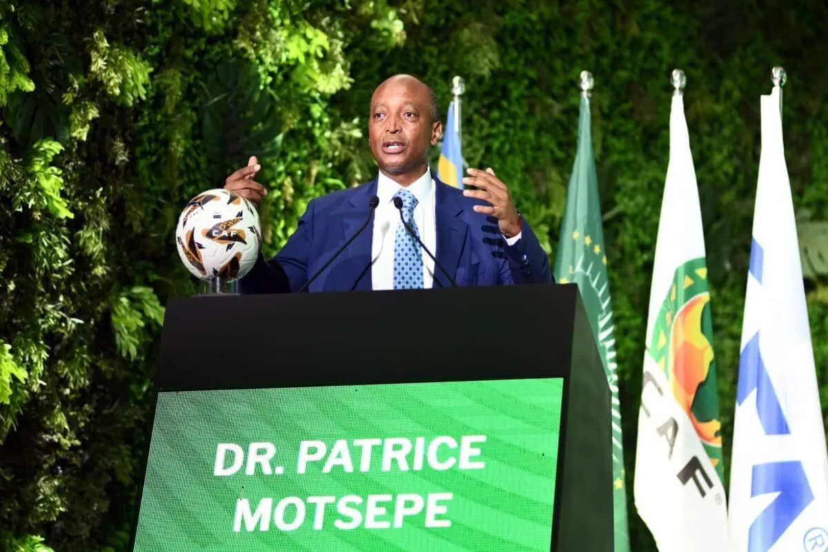 Man in a blue suit gives a speech at a podium with a CAF-branded soccer ball on the podium; flags stand behind him.