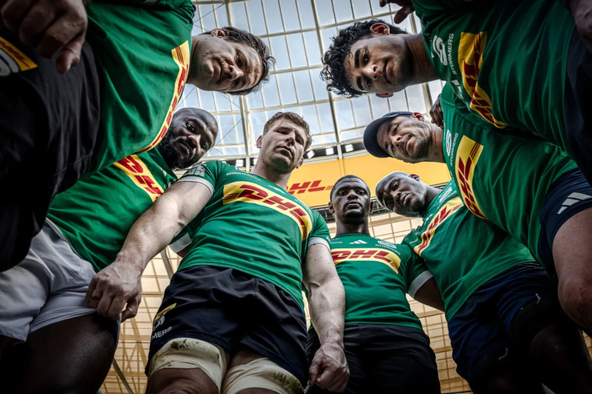 Rugby players in green DHL jerseys form a circle, looking down at the camera from a low angle in a bright indoor stadium.