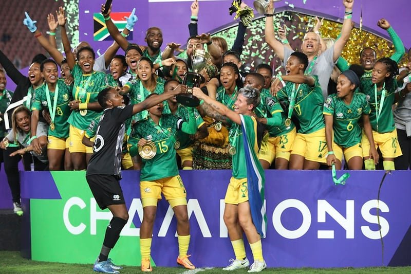 South African women's soccer team celebrating a championship, lifting the trophy as confetti falls behind them.