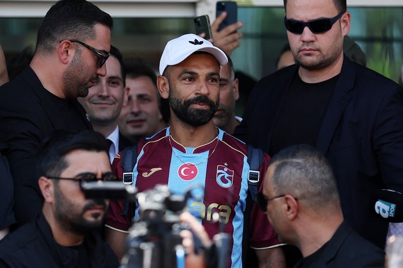 Bearded footballer in maroon-and-light-blue jersey and white cap surrounded by security and reporters photographed by cameras.