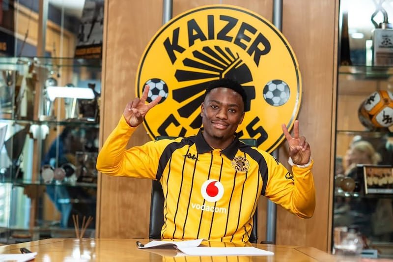 Young man in a yellow-and-black Kaizer Chiefs jersey sits at a table in a club office, flashing peace signs with both hands in front of a large Kaizer Chiefs logo.