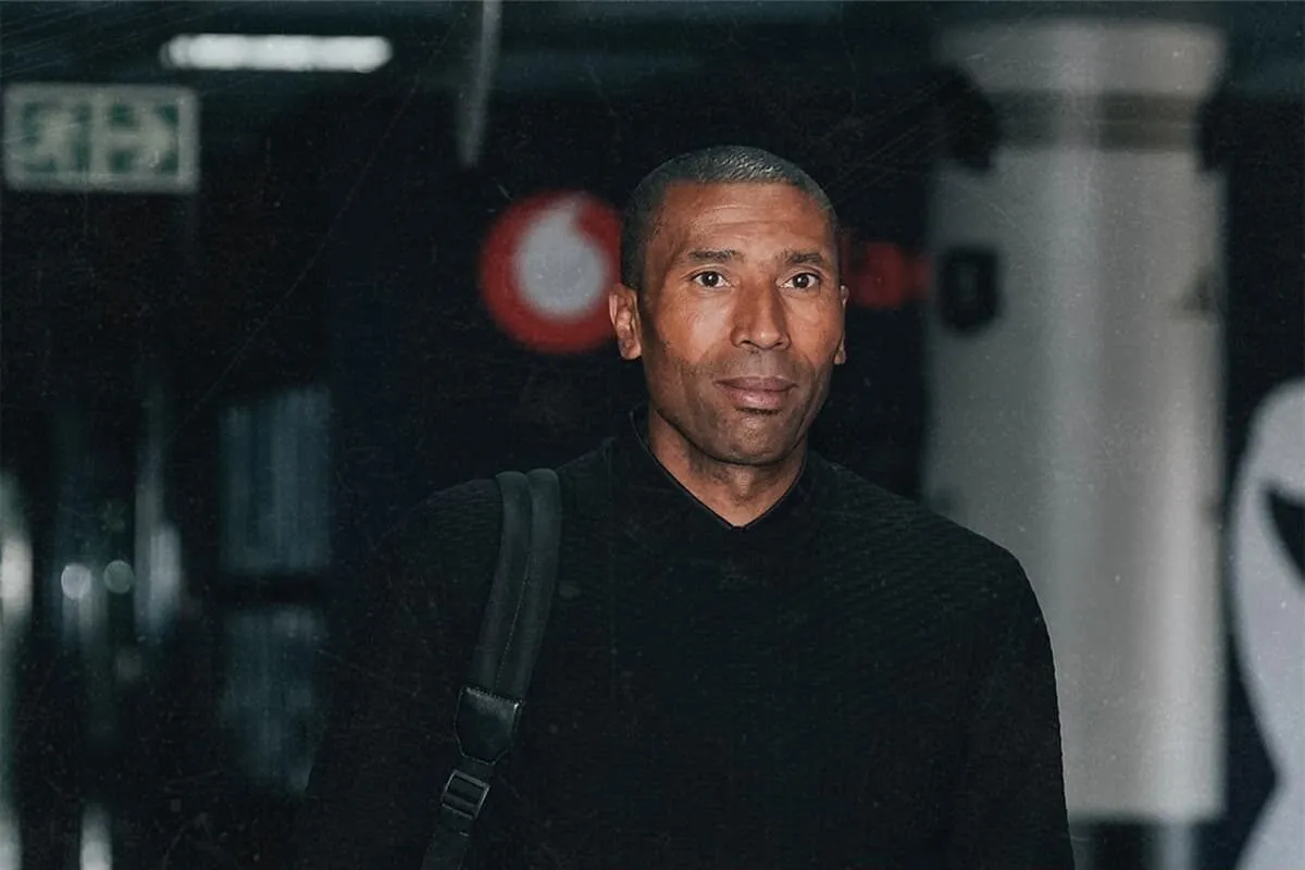 A man wearing a black turtleneck sweater with a shoulder bag stands in a dim indoor corridor.