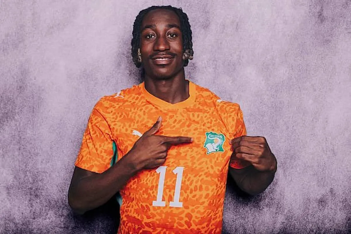 Athlete in an orange patterned jersey with number 11, pointing to the team crest on his chest against a purple backdrop.