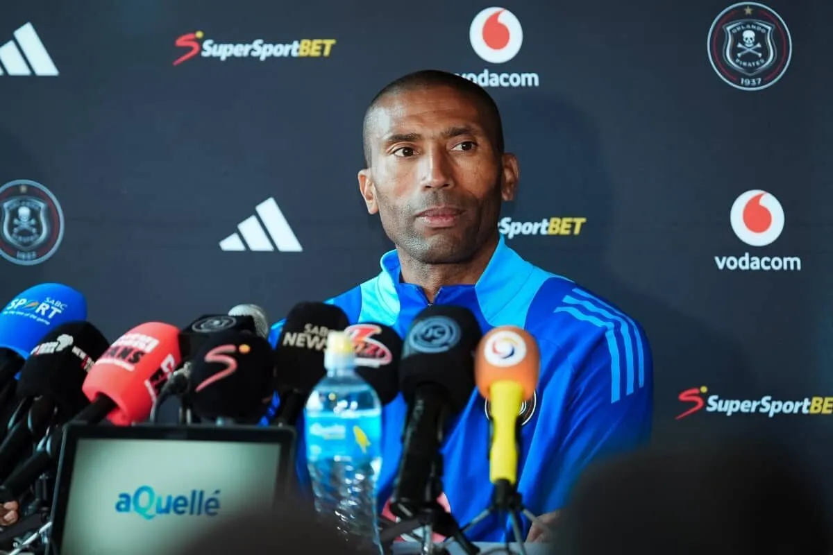 Man in a blue sports jacket at a press conference, surrounded by multiple microphones and media outlets on a dark backdrop with sports logos nearby.